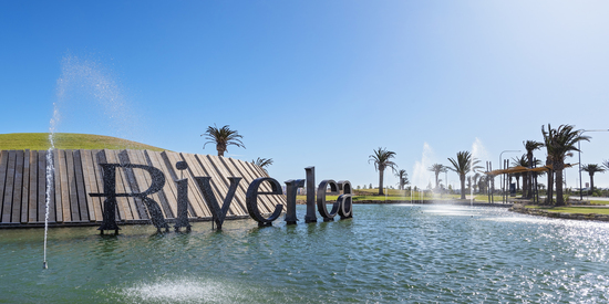 The Riverlea landmark sign at the entrance rises from a sparkling lake feature surrounded by palm trees, fountains, and manicured green lawns under a clear blue sky.