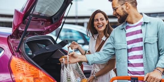 A smiling couple loads reusable mesh shopping bags into the open trunk of a pink car in a store parking lot.