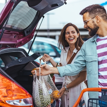 A smiling couple loads reusable mesh shopping bags into the open trunk of a pink car in a store parking lot.