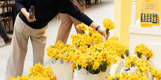 A shopper selects a bunch of bright yellow daffodils from white buckets at a charity flower stall inside a shopping centre.