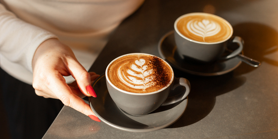 A woman’s hand reaches for a beautifully presented cappuccino with intricate latte art, alongside a second coffee on a cafe counter.